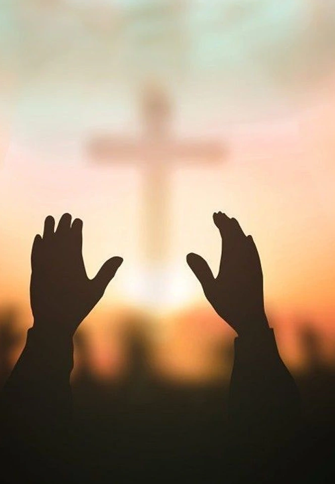 Hands lifted up in worship against a warm, glowing sunset with a soft-focus cross in the background.